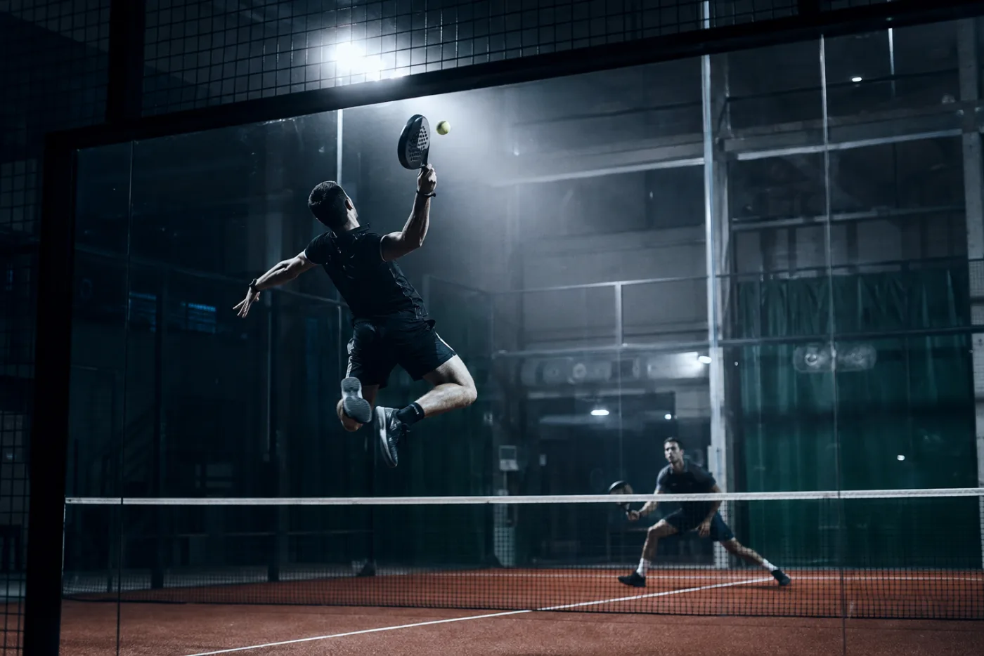 Spieler beim Sprungschlag am Netz, aufgenommen hinter der Glaswand des Padel-Courts